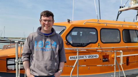 Karl Williams in a jumper with RNLI printed on it, stood in front of a large orange lifeboat. The boat has the nameplate reading RNLB Eric's Legend.