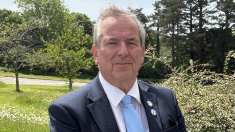 A man with grey hair wearing a dark blue suit with white tie and turquoise tie standing in front of some bushes and trees. 
