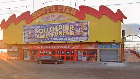 The entrance to South Pier, a large rectangular building painted yellow and red. It has a huge sign saying South Pier in yellow under a red roof shaped like a wave and many large red glass entrance doors with a small shop either side