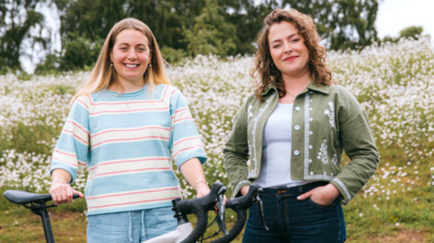 Two women stand in front of a field of white flowers. The one on the left has a pale blue jumper with red and white stripes and is holding a bike in front of her.

The woman on the right is in a green top over a white T-shirt, with her hands in her jeans pockets.  