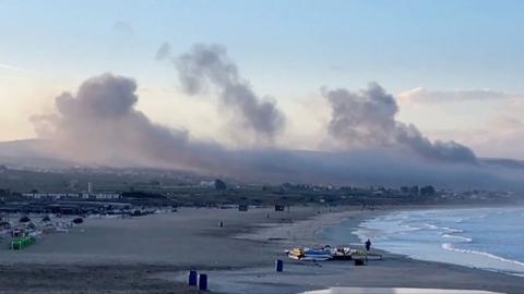 Smoke rises in the distance over a long beach