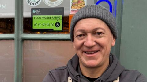 Marc Ward stands on the right of the image, looking towards the camera and smiling. He is wearing a grey hat and hoodie. He is standing in front of a building next to a window displaying a five-star food hygiene rating sign.