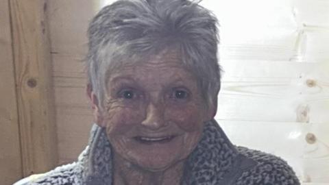 Joyce Crooks is smiling at the camera. She is wearing a grey fleece and has grey, short styled hair.
