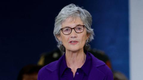Catherine Connolly, with short grey hair, wearing a purple top and purple jacket. She is standing behind a wooden podium. 