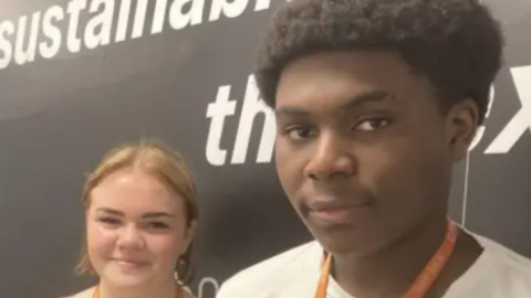 A woman with blonde hair, a light brown cardigan and an orange lanyard alongside a man with black hair a white T-shirt and an orange lanyard, standing in front of a black wall with a picture of a person's internal organs underneath a blue outline