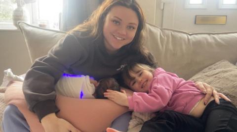A woman sat on a cream sofa with a baby wrapped in a blanket emitting blue light. A child with brown hair is laid on her mother with her hand on the baby's head. She is wearing a pink jumper.