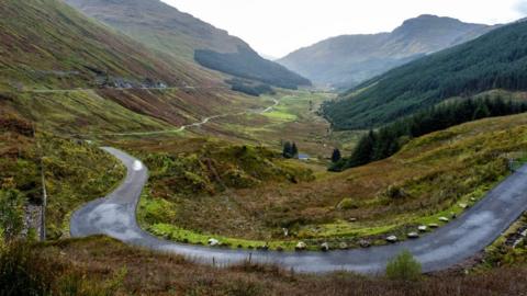 The Old Military Road beside A83 in Argyll at the Rest and Be Thankful. A tarmac road curves around the front the image and runs into the distance, below the main road. There are green hills on either side of the picture. There's a dense section of forest on the right. 