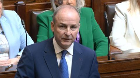 A man in a blue suit and blue tie stood speaking in the chamber