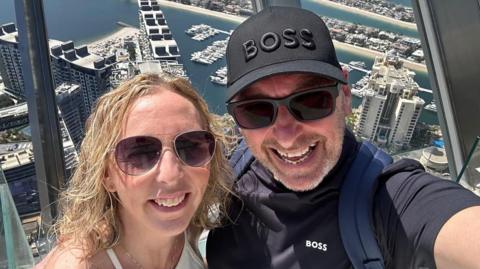 Cynnamon and Tony Church smile from the top of a building overlooking a marina in Dubai. Tony has a black Boss baseball cap on and is wearing a dark Boss T-shirt with a rucksack on his back. Cynnamon, who has curly blonde hair, is wearing a white top. Both are wearing sunglasses.
