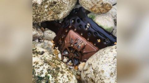 The brown hatch door of a submarine wreck attached to a black metal outer piece of the wreck with holes in it. The hatch door piece is wedged amongst large and small stones