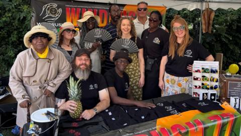 Members of the Windrush society sit on a stall selling T shirts, smiling and holding various items including fans and a pineapple. Many are wearing Windrush society t-shirts.