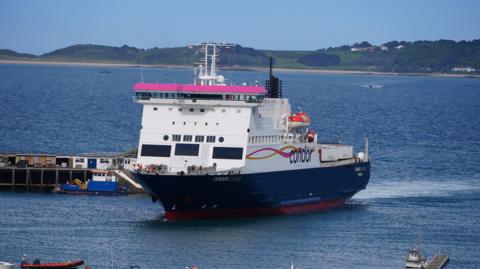 A blue and white Condor ferry with pink and yellow branding on its port side. It is sailing into harbour and there is a rigid inflatable boat in the foreground. The mainland can be seen over the water behind the ferry.