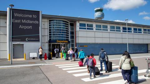 A photograph of the outside of East Midlands Airport. You can see the door and a sign saying "Welcome to East Midlands Airport" on the left. People are crossing the road in front of the airport and walking towards the building 