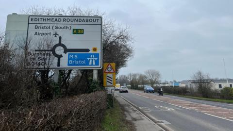 A road and sign with several cars on it driving past. There's a caravan park to the right. The main shot is of the roundabout entrance sign. It says Edithmead Roundabout with a left turn to Burnham-on-Sea, straight on to Bristol Airport and right to the M5 towards Bristol.