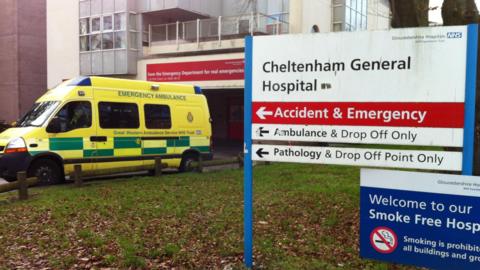 A sign for Cheltenham General Hospital Accident and Emergency department. The sign also says 'Ambulance & Drop Off Only' and 'Pathology & Drop Off Point Only'. There's another sign below it saying 'Welcome to our Smoke Free hospital'. Behind the signs is an ambulance parked in front of a hospital building. 