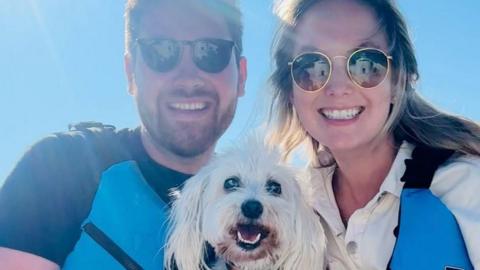 Joe (left) and Charlotte (right) smile at the camera with Max in the centre. They are all smiling at the camera. Joe and Charlotte are both wearing sunglasses and blue lifejacket vests. 