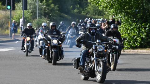 A large convoy of motorcyclists driving down the road, with puffs of smoke coming from their exhausts. 