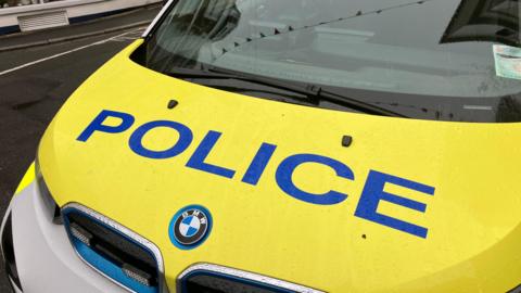 The image shows a BMW police car with a yellow bonnet with blue police writing across it. There is a reflection of bunting in the windscreen and the car is positioned on an unknown Guernsey street in damp conditions.