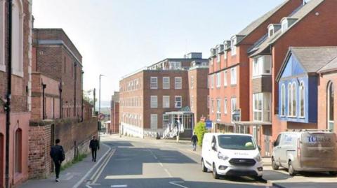 Buildings can be seen on either side going down this road, with four people seen walking and two vans parked outside a building on the right hand side.