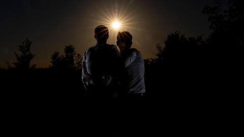 Jerry and Jamie are in silhouette as the moon clouds the sun. They each have an  arm around each other as they gaze towards the eclipse. 