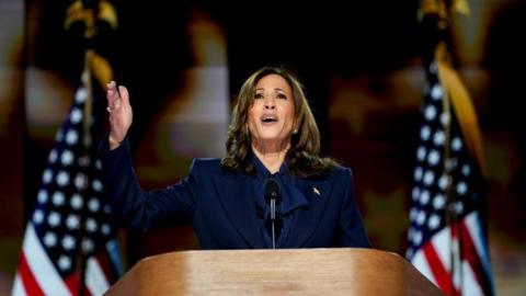 Kamala Harris speaking and gesturing at a podium, with American flags behind her