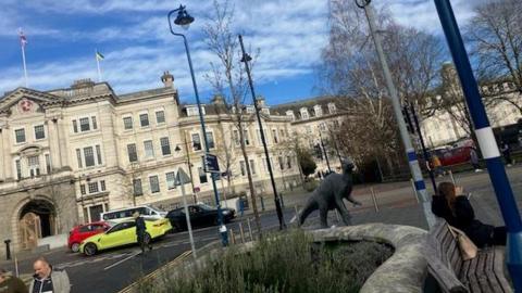 The image shows Kent County Council in the background, with cars driving past and people walking on the pavements around it. A girl can be seen on the right-hand-side sat on a bench, which is in front of a small plot of grass.