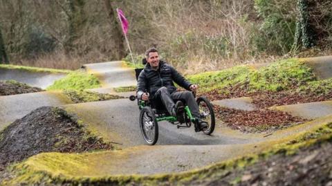 A man on a three wheeled tricycle with a pink flag on the back pedalling along wavy tracks at the new green space. He is wearing black jeans and a black padded coat and is smiling