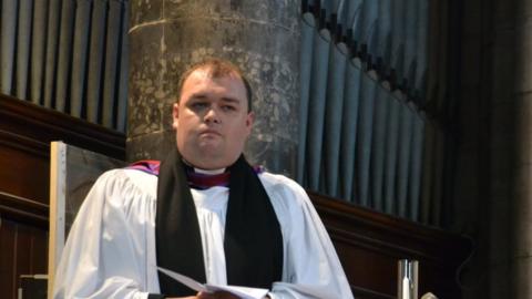 Samuel Erlandson in white and black robes. He is holding a piece of paper and standing in front of an pipe organ