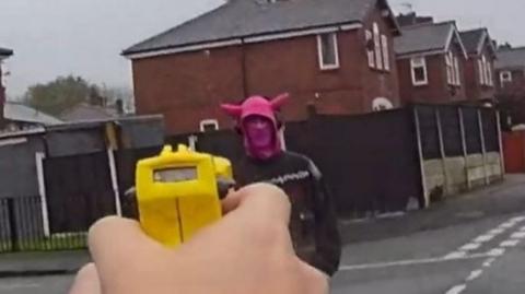 A still from police bodycam footage showing the moment the PC holds up a Taser to dispatch it at the suspected armed robber, who is stood facing the PC wearing dark clothing, a pink mask or scarf across the lower part of his face, and a pink hat with two pink horns sticking out of it. There are residential red brick terraced houses in the background and the sky is grey.