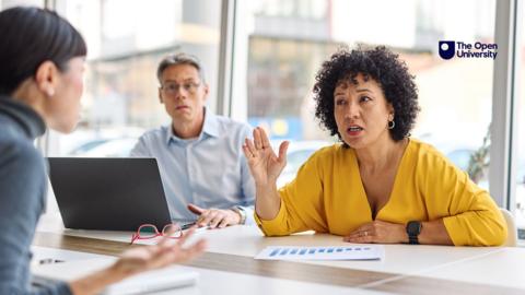 Two colleagues argue at a desk while another watches on in the background