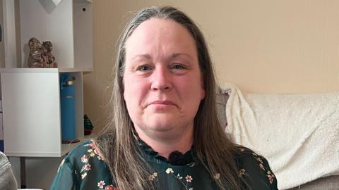 A woman looking at the camera, she has dark blonde hair