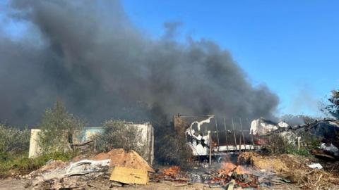 A pile of burning rubbish and sheds surrounded by thick black smoke which is filling the sky