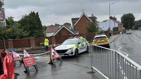 Roadworks signs are next to a road which has also been cordoned off by police, with two cars blocking the road and four officers are stood nearby