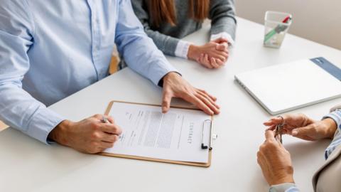 Three people sitting around a white table. None of their faces are shown. One is holding a set of keys, another, wearing a blue shirt, is signing a contract. A third person at the top of the image is wearing a grey top over a white shirt and has her hands clasped. 
