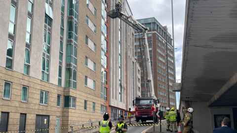Police and fire officers stood on a street that is taped off, as a fire engine with a hydraulic platform is parked next to high-rise buildings. Two fire officers are on the platform inspecting a building. More buildings can be seen in the background.