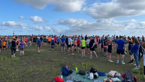 A large group of people are dressed in running gear in a huge field. They are standing around and some are chatting.