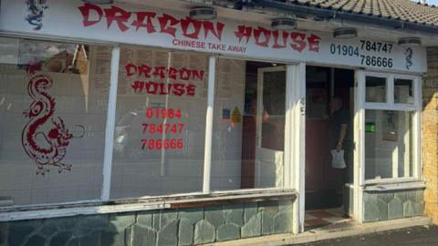 A Chinese takeaway called Dragon House which has been cleaned after being covered in graffiti. It has large windows with red writing on and a picture of a dragon. 