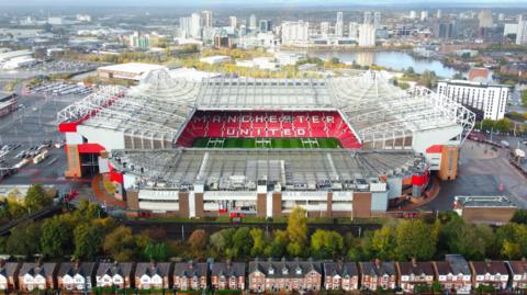 An aerial photograph taken by drone of Old Trafford stadium, which can be seen closed to a row of semi-detached houses, surrounded by pedestrian areas and car parks. In the distance, high-rise buildings can be seen on Salford Quays.