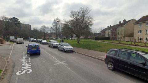 Sweetbrier Lane in Exeter with several cars parked alongside the pavement on one side of the road and with vehicles queueing at traffic lights.