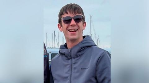 Hayden Davies smiles at the camera wearing sunglasses wearing normal civilian clothes in a photo taken before he was in Russian captivity. 