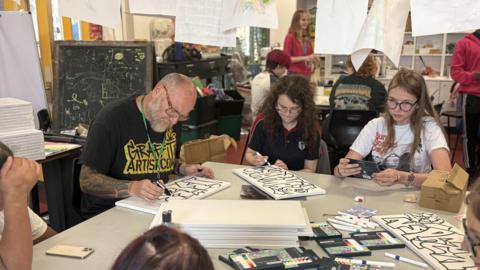 Men and women are sitting on a table holding pens and drawing fonts on canvases. They are sitting in an art room with work drying from the ceilings.