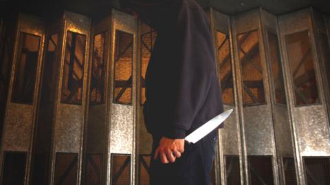 A man in a dark blue hoody stands side on to the camera holding a large kitchen knife in his left hand.