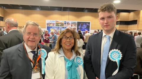 Three people wearing “Reform UK” rosettes stand together in a busy indoor hall, dressed in formal attire. Behind them, groups of attendees gather and converse near a stage area with posters, under bright overhead lighting and a high, wood-paneled interior.
