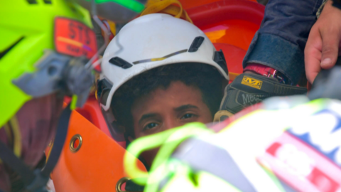 the picture shows half of the boys face - the rest is obscured by rescue workers. He's wearing a white hard hat and has dark curly hair.
