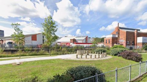 A green fenced area is in front of grey metal factories and brick buildings.