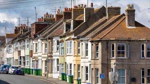 Terraced houses of a variety of colours. There are green bins out the front of the houses, and a number of cars parked on the road. 