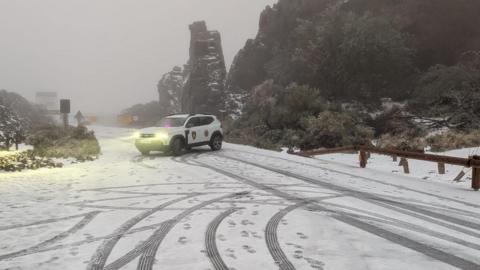 A police car with its headlights on sits across a road covered by a thin later of snow, with tyre-tracks, greenery in the background and a grey murky sky overhead