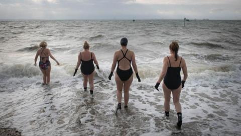 Four swimmers in black wetsuits and gloves walk into the sea as the waves crash in. The sky is grey.