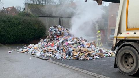 The pile of rubbish dumped in a pub car park. The rubbish is smouldering. A firefighter is training a hose on it. The back part of the bin lorry is also in the picture