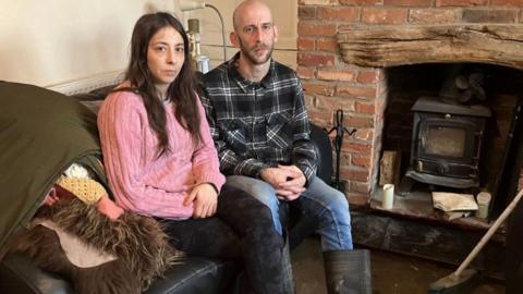 Tom and Jade sit on a sofa near to a wood burning stove. Jade is wearing a pink jumper and black trousers. Tom is wearing a check shirt, jeans and a pair of wellies.